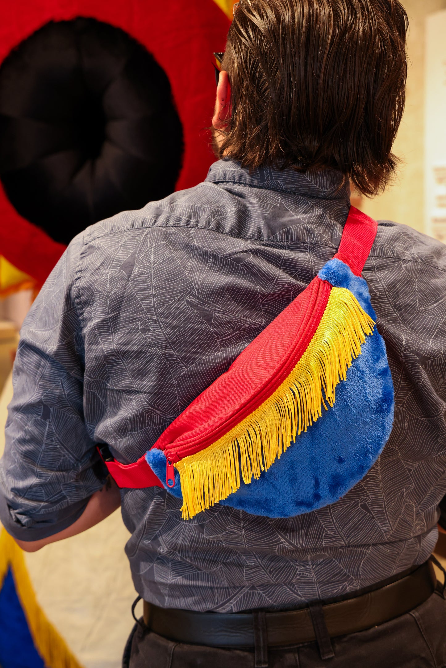 A person with shoulder-length hair and a blue patterned shirt carries a Fringe Fanny Pack - Yellow over their shoulder. They face artwork indoors, exuding trendy style similar to that of stylish fringe fanny packs.