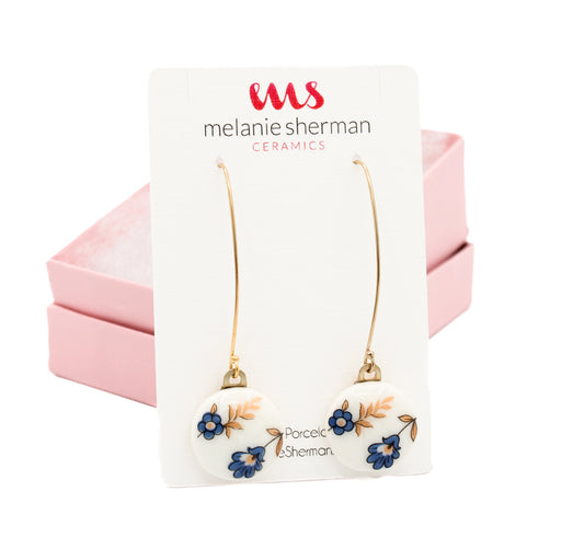 Gold-filled round porcelain dangle earrings with vintage blue flower designs are displayed against a card labeled Melanie Sherman Ceramics, beautifully nestled in a pink box. Product Name: Porcelain Dangle Earrings Blue 2 Flowers.