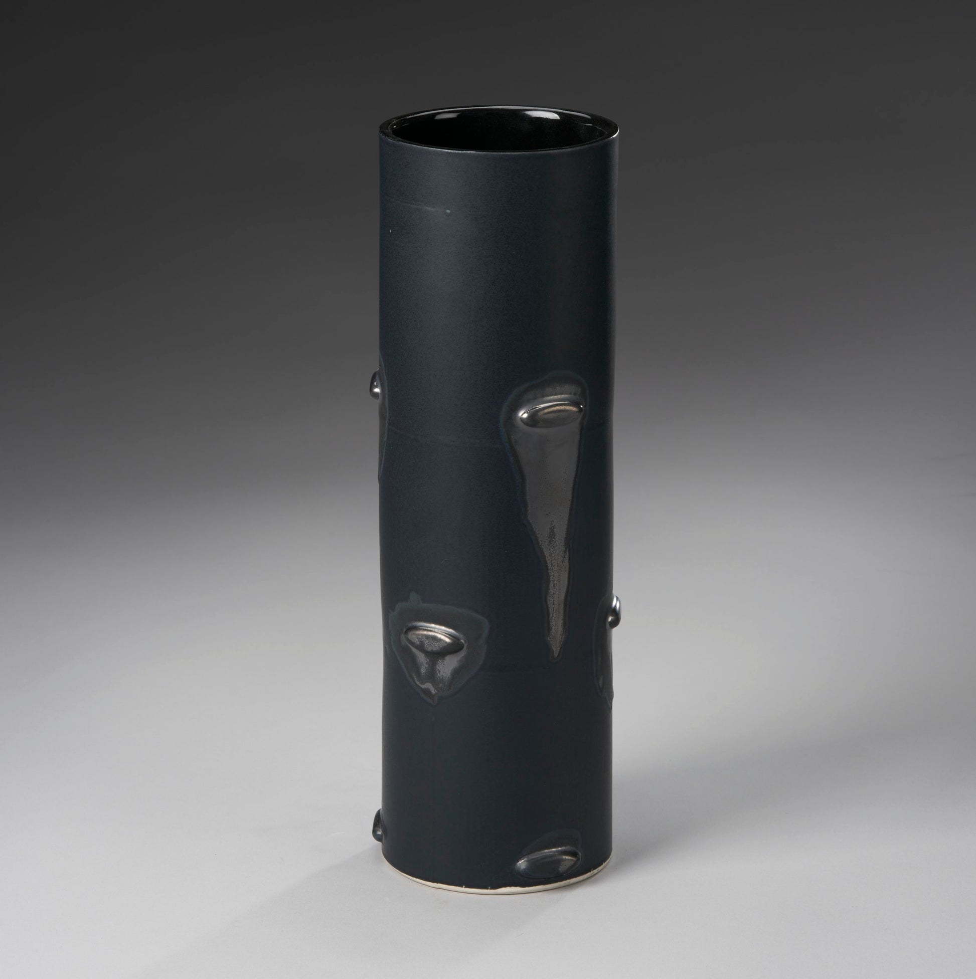 The Large Vase by Mikey Gambino is a tall, smooth, dark ceramic piece with a cylindrical shape and glossy drips. Set against a gradient gray backdrop, its sleek design highlights masterful slip casting techniques by the skilled ceramic artist.