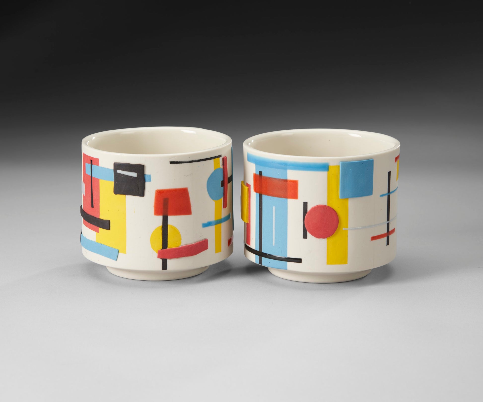 The Colorful Lovers set by Mikey Gambino features two slip-cast ceramic tumblers with a modern abstract design. Geometric shapes in red, blue, yellow, and black adorn the white background, creating a dynamic pattern with rectangles, circles, and lines.