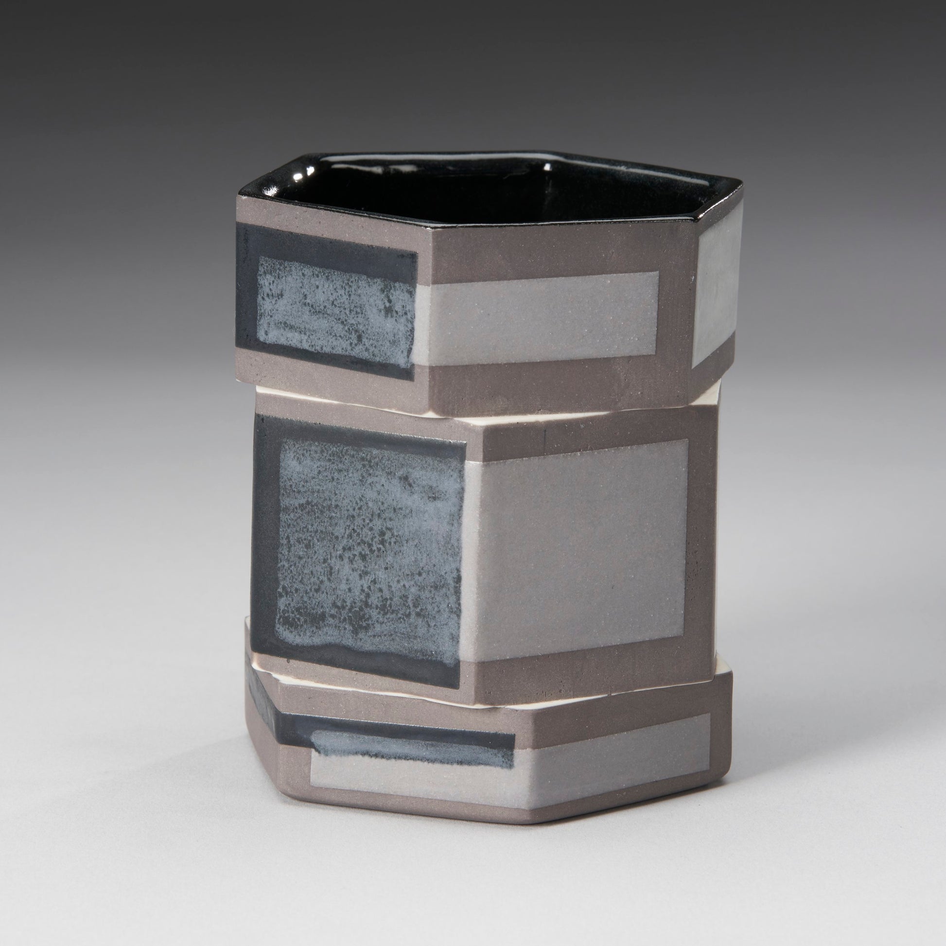 The Small Twisted Hexagon Mug by Mikey Gambino features a geometric hexagonal design in shades of gray. With a blend of matte and glossy finishes and rectangular patterns crafted using slip casting techniques, it showcases a modern, artistic flair.
