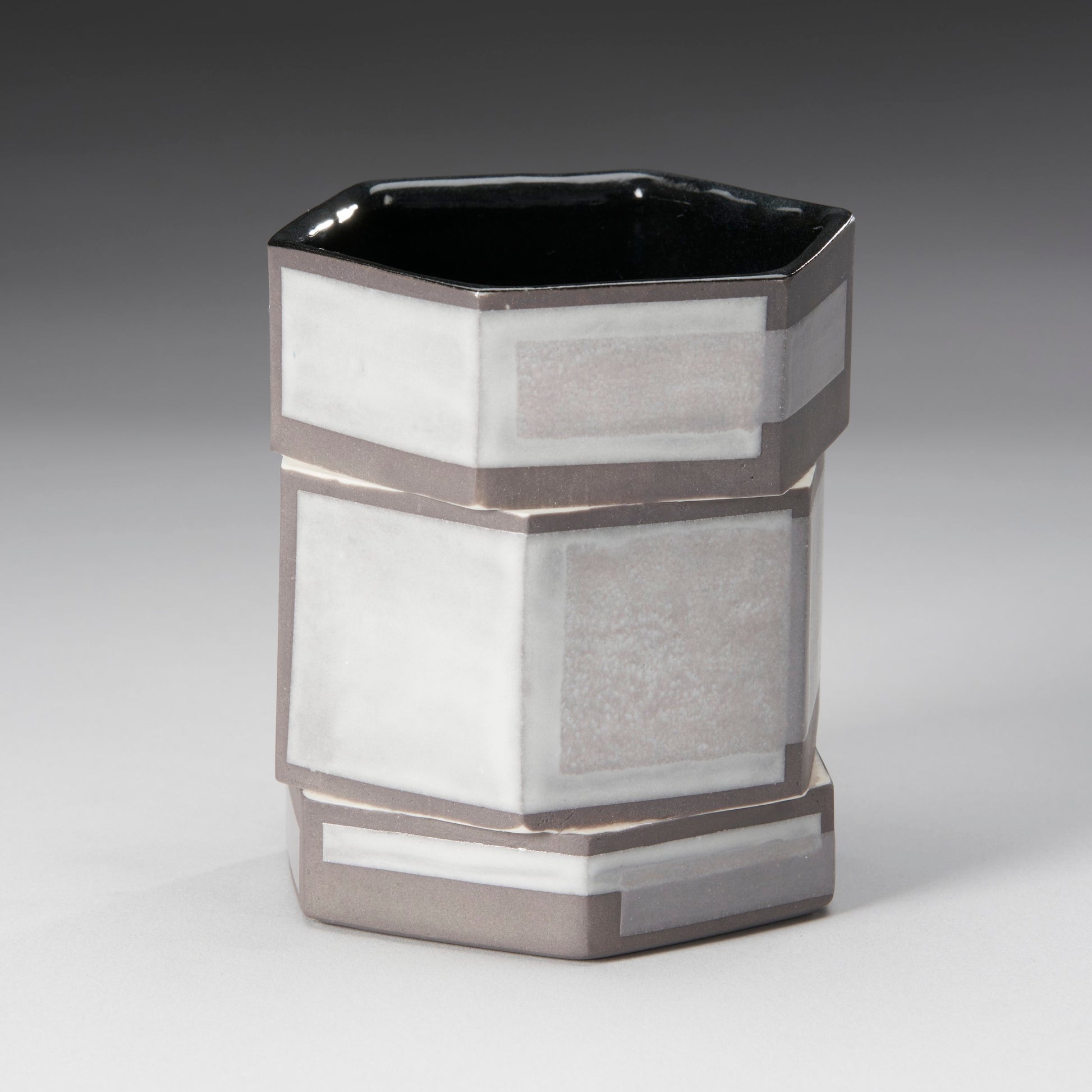 The Small Twisted Hexagon Mug by Mikey Gambino is a glossy ceramic cup with a skilled artisans touch, featuring alternating white and gray patterns on each side. Its dark interior contrasts elegantly with the light exterior, offering a modern geometric flair.