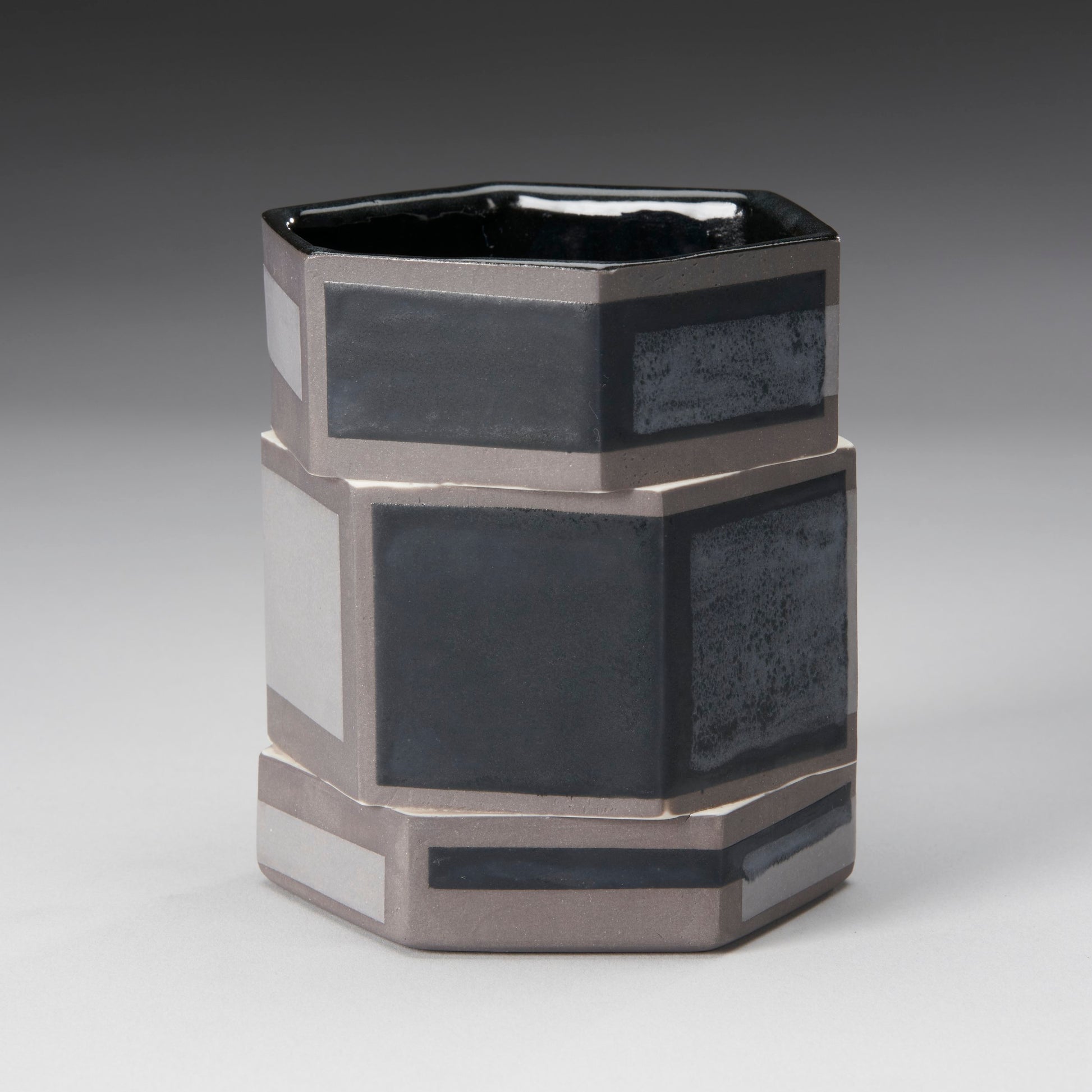 The Small Twisted Hexagon Mug by Mikey Gambino is slip-cast with a modern geometric design, featuring alternating black and gray rectangles. This ceramic artists piece has a smooth, glossy finish set against a neutral gradient background.
