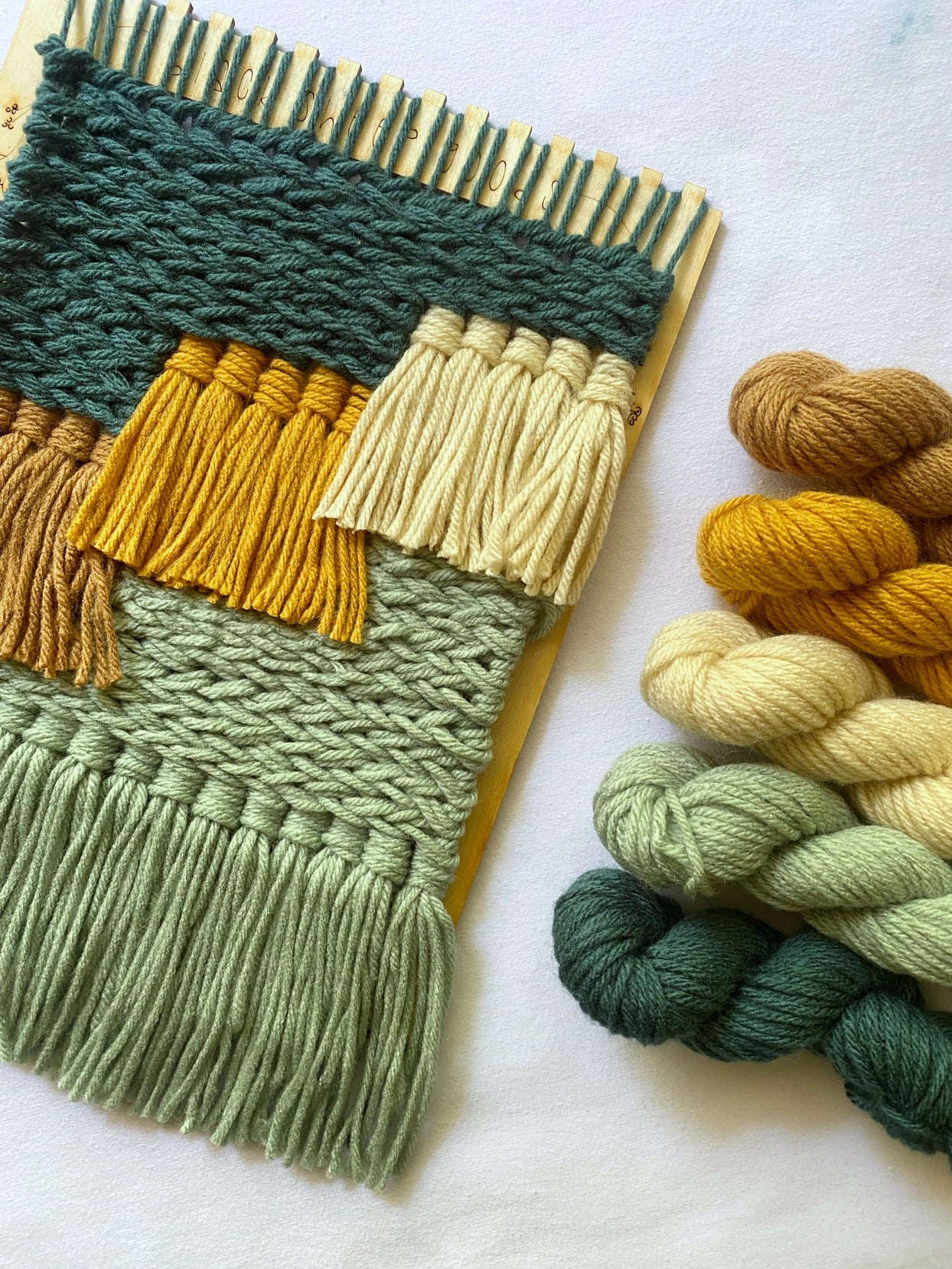 The Tapestry Weaving Kit Woodlands offers a woven wall hanging in green, mustard, and cream yarn on a wooden loom. It includes matching yarn skeins and features textures with fringes at the bottom, ideal for anyone passionate about detailed craftsmanship.