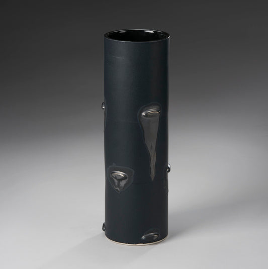 The Large Vase by Mikey Gambino is a tall, smooth, dark ceramic piece with a cylindrical shape and glossy drips. Set against a gradient gray backdrop, its sleek design highlights masterful slip casting techniques by the skilled ceramic artist.